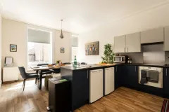 A bright, modern kitchen with dark blue and cream cabinets, wooden flooring, a central island, dining table and chairs by two windows, integrated appliances, a microwave, houseplants and colourful wall artwork. A black recycling bin sits beside the island.