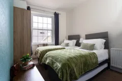A double bedroom with white bedding, a green throw, bedside tables, lamps and a window overlooking the street.
