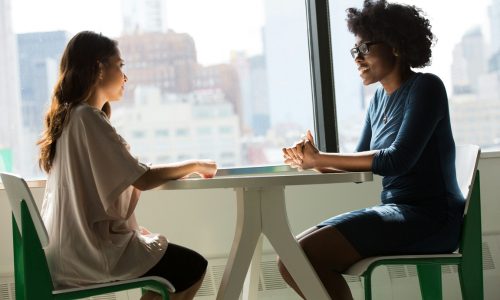 A photo of two young women sat opposite each other at a table by a window.