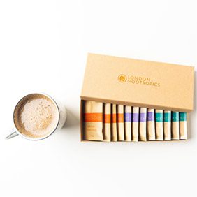 Open box of London Nootropics coffee blends showing colorful sachets, next to a cup of creamy coffee on a white background.