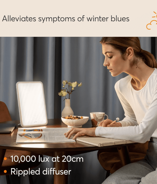 A woman using the Lumie light therapy lamp at a desk, emitting 10,000 lux brightness.