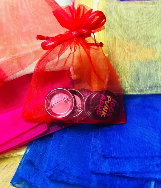 A small red organza gift bag filled with several button badges, sitting on a vibrant, multicolored fabric background.