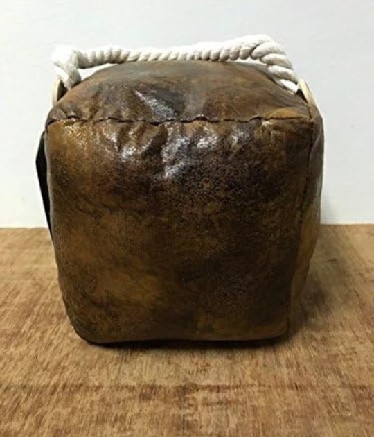 A brown leather doorstop in a cube shape with a rope handle, placed on a wooden surface.
