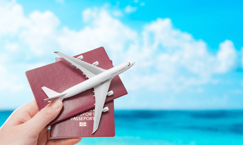 Hand holding two red passports with a toy airplane placed on top, against a bright blue sea and sky background.