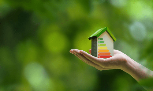 Hand holding a miniature house with an energy efficiency rating chart displayed on its side, against a blurred green background.