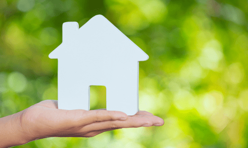Hand holding a white cutout of a house against a lush green natural background, symbolizing honest sustainable housing.