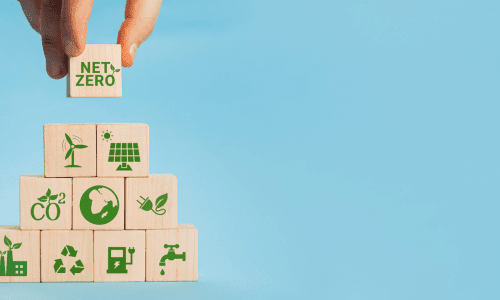 Wooden blocks stacked in a pyramid with eco-friendly icons such as renewable energy, water conservation, recycling, and carbon neutrality, topped with a block labeled ‘Net Zero.’