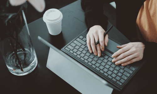 Close-up of hands typing on a laptop keyboard with a stylus, coffee cup nearby, and a vase with flowers in the background.