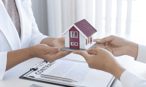 Two people holding a miniature model house above a contract on a table with a pen and currency visible, symbolizing property agreements and landlord support.