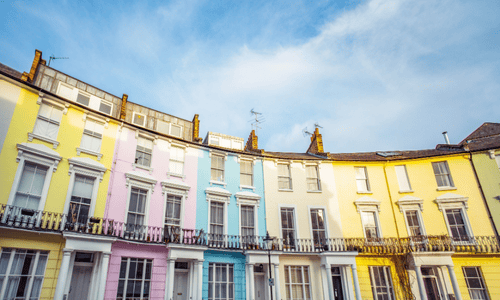 Pastel-colored terraced houses with tall windows and ornate balconies, lit by the afternoon sun against a blue sky.