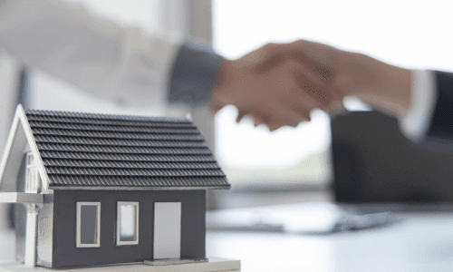 Model house in the foreground with two people shaking hands in the blurred background, symbolizing rental agreements.