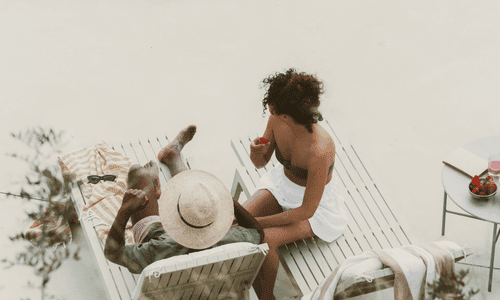 Couple relaxing on lounge chairs, one wearing a straw hat and the other in a bikini, sharing strawberries by a small side table outdoors.