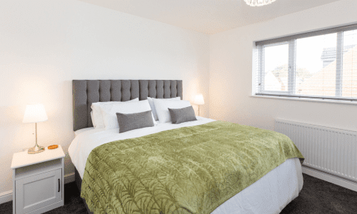 Modern bedroom with a large bed, grey upholstered headboard, white linens, and a green patterned throw blanket, with bedside lamps and natural light.
