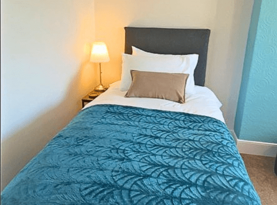 Compact bedroom with a single bed dressed in white linens and a teal throw, positioned beside a window with natural daylight, featuring a bedside table with lamp, soft carpet flooring, and a wardrobe in a bright, simple layout.