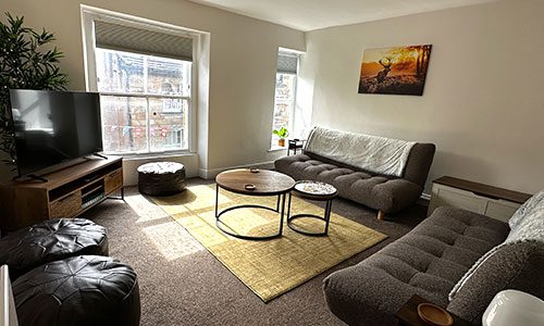 A cosy living room photograph with sun blasting through the 2 large windows in the back and 2 large sofas facing a TV in the corner.